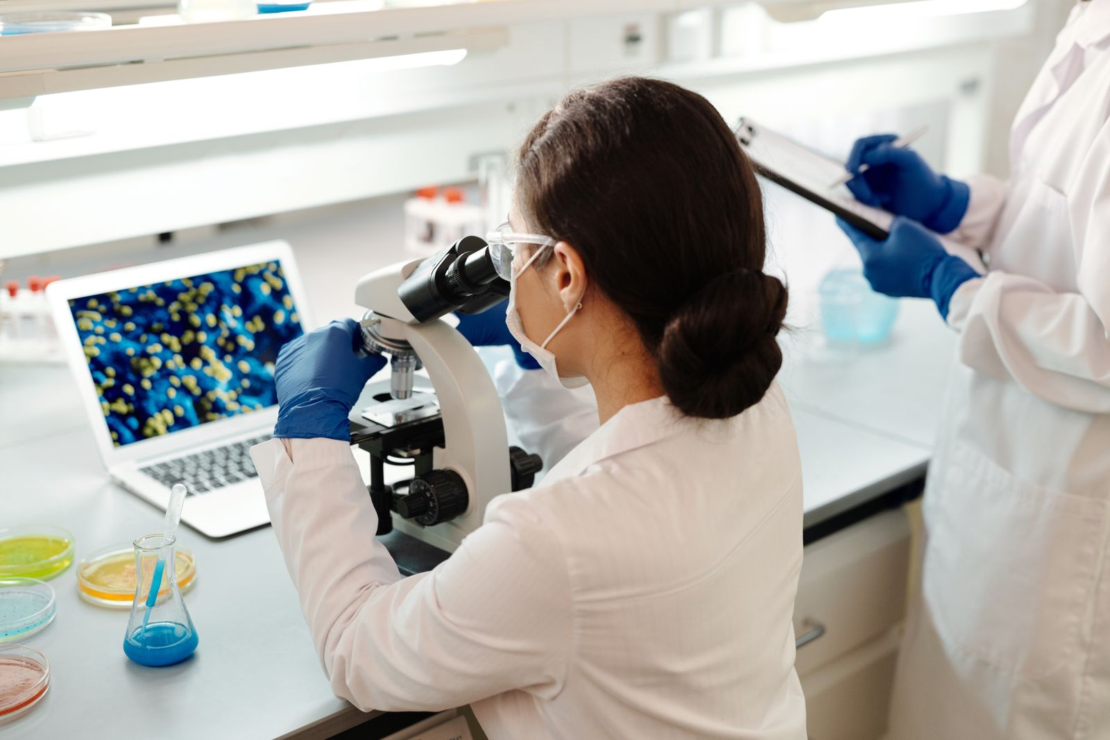 Two pathology scientists in a laboratory, one at a microscope and one holding a clipboard